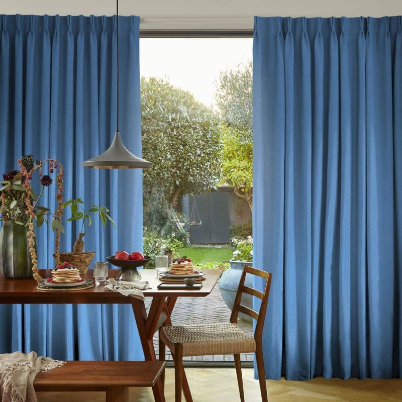 Blue pleated curtains, drawn partly open to sides, framing and filtering sunlight; a dining table set for breakfast with pancakes and pendant lamp, dining room opening to a leafy backyard.