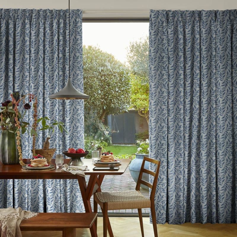 Blue-and-white leaf-patterned full-length curtain panels frame a central opening, partially drawn to reveal a sunlit garden; wooden dining table set with pancakes and a pendant lamp in a cozy breakfast nook.