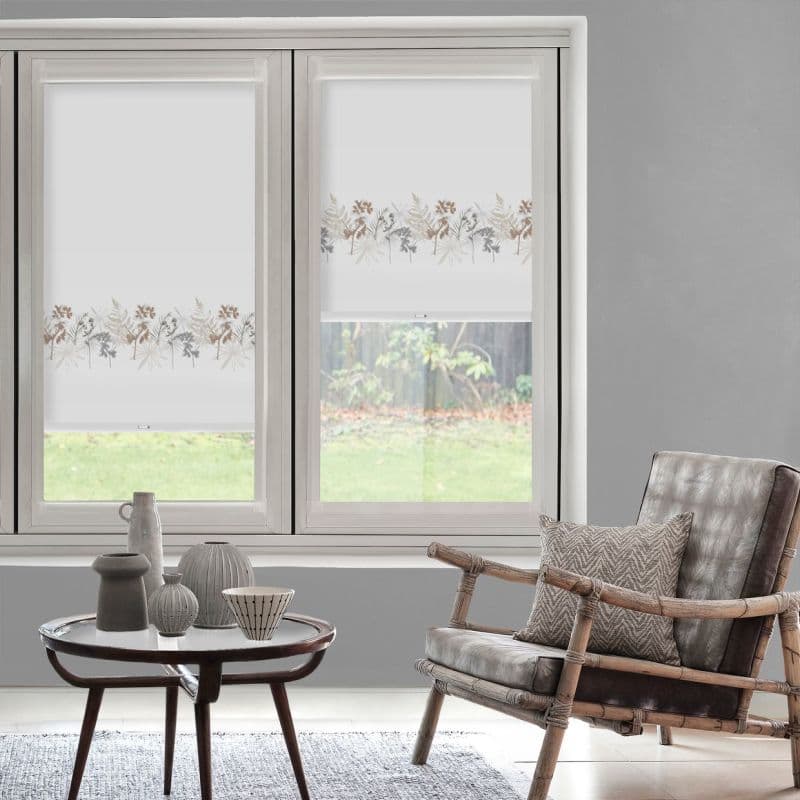 White roller blinds with a horizontal watercolor floral band across mid‑height, partially lowered, filtering soft daylight in a neutral living room with a rattan armchair, coffee table, and garden view.