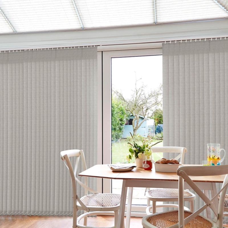 Light-gray fabric vertical blinds on a ceiling-mounted track, drawn closed with a central gap; filtering daylight through patio doors, overlooking a small garden; dining table and chairs in foreground.