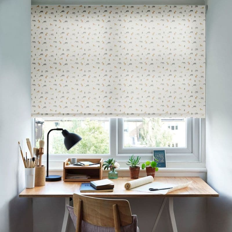 Roller blind with repeating animal print, fully lowered over a window, filtering soft daylight; wooden desk beneath holds a lamp, art brushes, notebooks and potted succulents, view beyond.