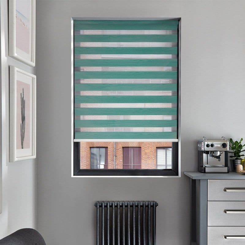Teal zebra roller blind, lowered mid-window, with alternating opaque and sheer horizontal bands filtering daylight; set in a gray kitchen room with radiator beneath, countertop espresso machine and cactus prints.