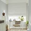 White textured Roman shade lowered over a window, blocking most light and letting soft daylight from the bottom; small home office desk with wicker chair, lamp and potted plants. 01