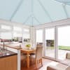 Pleated pale-blue roof blinds cover the conservatory ceiling, closed and diffusing light; white pleated window and door blinds partly lowered; bright dining-kitchen with wooden table, chairs and patio view.