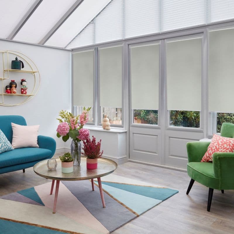 Light-colored roller blinds, lowered mid-height across tall conservatory windows, partially filtering daylight and leaving bottom panes exposed; modern sunroom with pastel furnishings, low coffee table and garden glimpses outside.