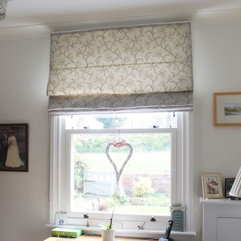Cream fabric Roman blind with grey vine embroidery, lowered halfway and softly filtering daylight; hangs over a white-framed sash window in a bright room with photos, desk and garden view.