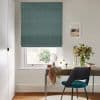 Teal textured Roman blind lowered mid-window, slightly above the sill, filtering daylight; wooden desk beneath with mirror, teal chair and vase of flowers — bright home office with a garden view outside.