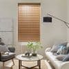 Wooden horizontal Venetian blinds, centered on the wall with narrow warm-toned slats, fully lowered and tilted to filter soft daylight; neutral living room with sofa, armchair, round coffee table, lamp.