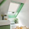 A beige pleated skylight blind, mounted inside an angled roof window, half-lowered, filtering daylight over a green-trimmed bathroom with white herringbone tiles, sink, bathtub, toiletries and a potted plant.