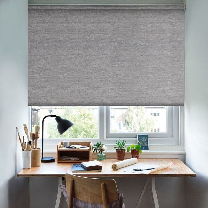 Grey woven roller blind lowered above the sill, softly filtering daylight over a wooden desk with lamp, brushes, notebooks and potted plants, against a daytime view of trees and nearby buildings.