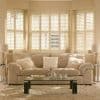 Cream-colored wooden plantation shutters covering a three-section bay window, slats partially angled to filter warm daylight; beige living room with plush sofa, fluffy cushions, glass coffee table and shag rug.