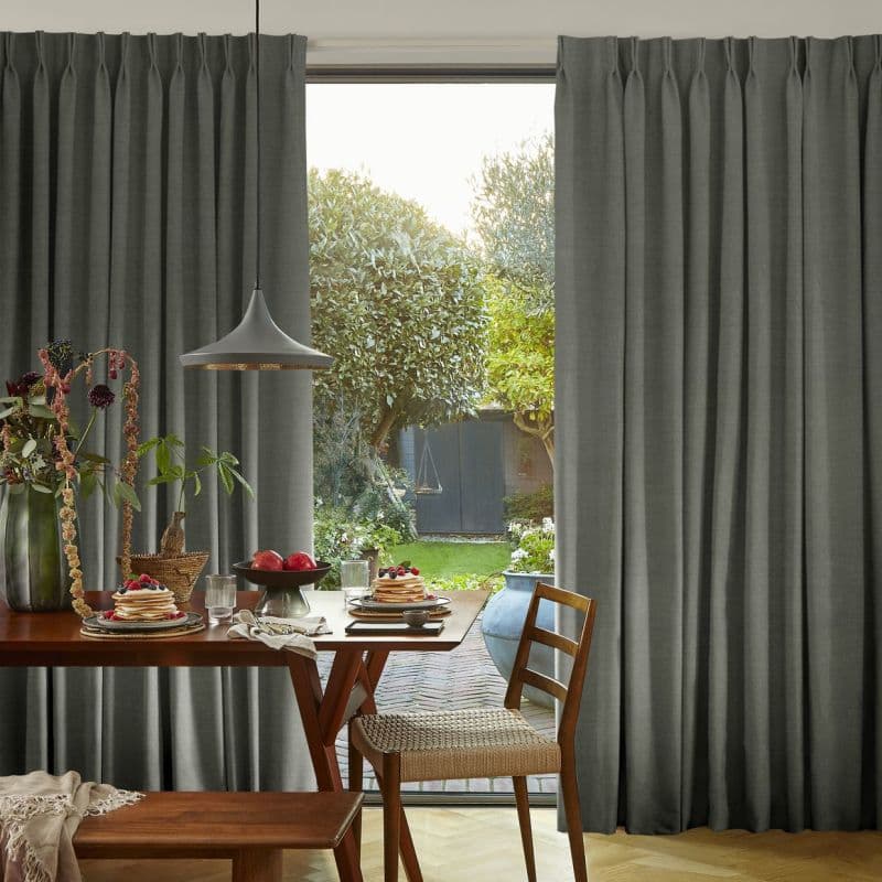 Floor-length pleated gray curtains, hung across a sliding glass door, parted at center; filtering soft daylight and framing a dining table set for breakfast and a leafy backyard view.