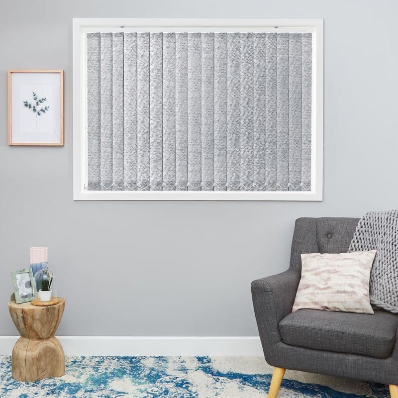 Vertical textured fabric blinds, closed across a white-framed window, lightly filtering daylight; set in a gray-walled living room with a gray armchair, knitted throw, wooden side table and blue rug.
