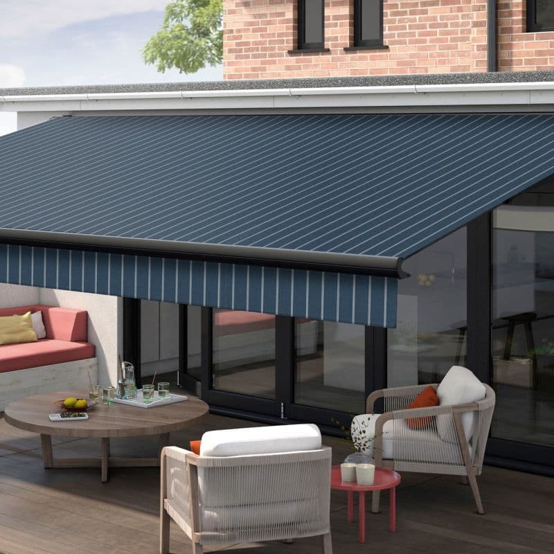 Blue striped retractable awning with matching valance extended over patio glass doors, shading a furnished terrace—wooden coffee table, cushioned chairs and bench—attached to a brick-faced modern house under daylight.