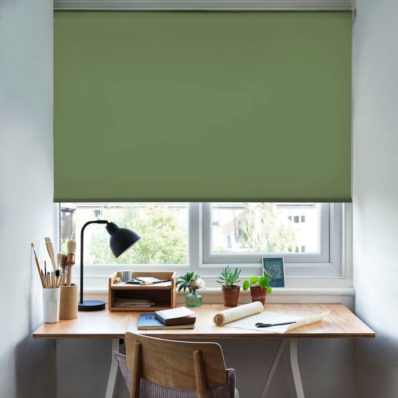 A green roller blind lowered across a desk-height window, mostly blocking light; a wooden desk beneath holds a lamp, brushes, notebooks and small potted plants, with daylight glimpsed below.