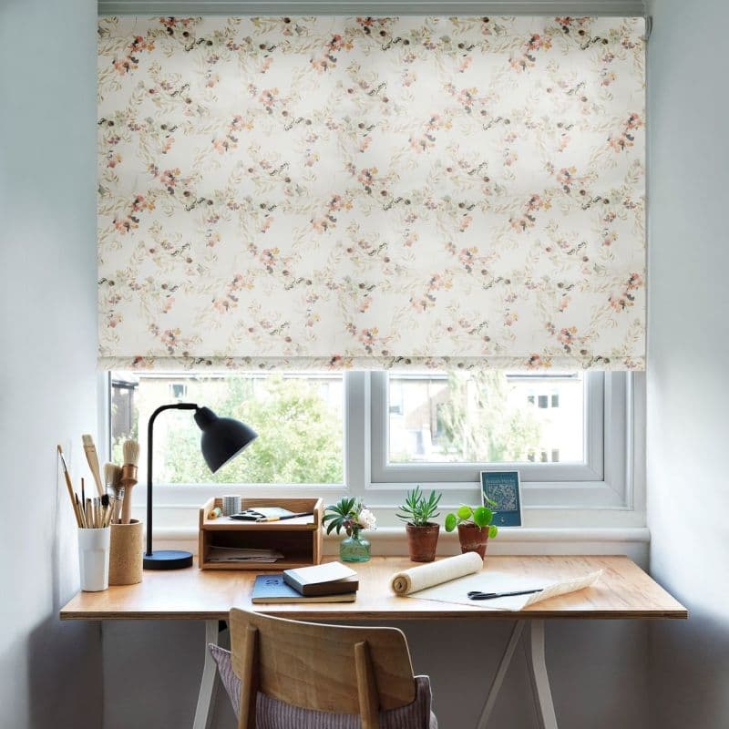 A cream roller blind with delicate peach-and-green floral print is lowered over a double window, filtering daylight above a wooden desk staged with a lamp, potted plants and art supplies.