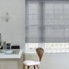 Sheer grey Roman blind, lowered to mid-window with soft horizontal folds, filtering diffused daylight; minimal home office with white desk, wooden chair, paper pendant lamp, and faint tree silhouettes outside.