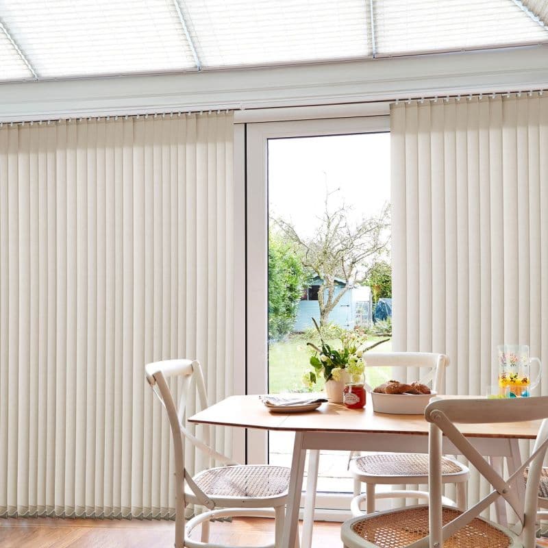 Vertical beige blinds, floor-to-ceiling on a headrail — parted at the center, filtering soft daylight — over a dining table and chairs in a bright conservatory, view of a garden with a tree and shed.
