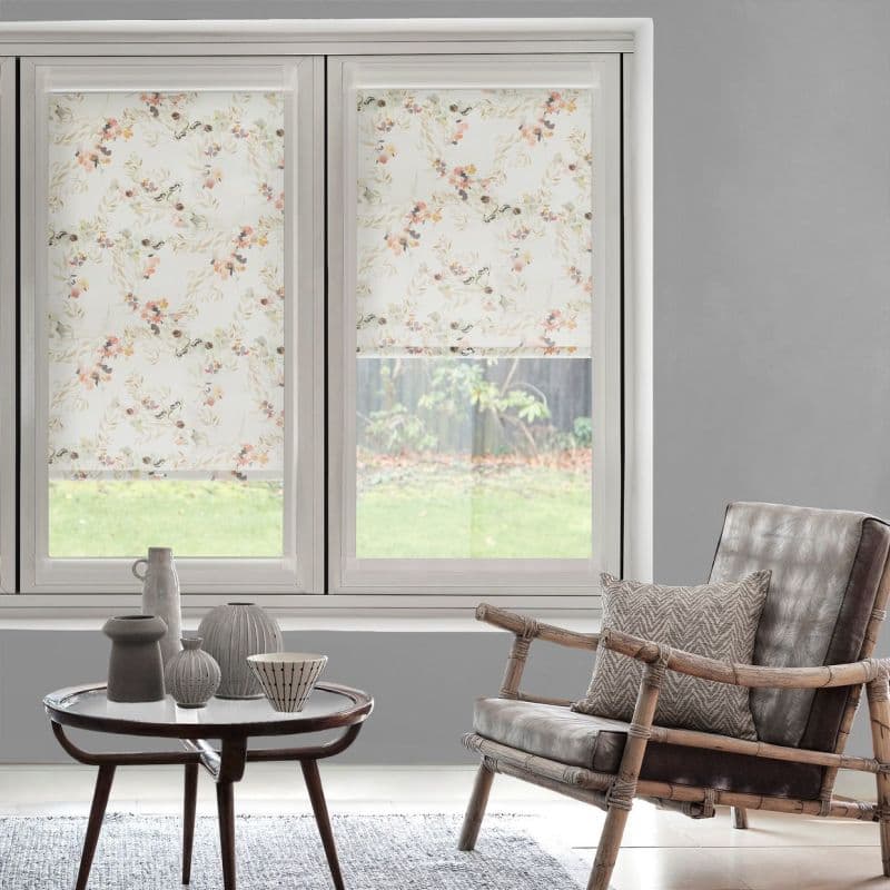Floral fabric roller blinds (two adjacent panels), pale botanical print, partially lowered and filtering daylight over a ground-level window; modern sitting area with gray walls, rattan armchair and small table.