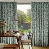 Botanical-print, floor-length curtains at a sliding glass door, parted to frame and let sunlight through; dining table with pancakes and vase beneath a pendant lamp opening onto a sunlit garden.