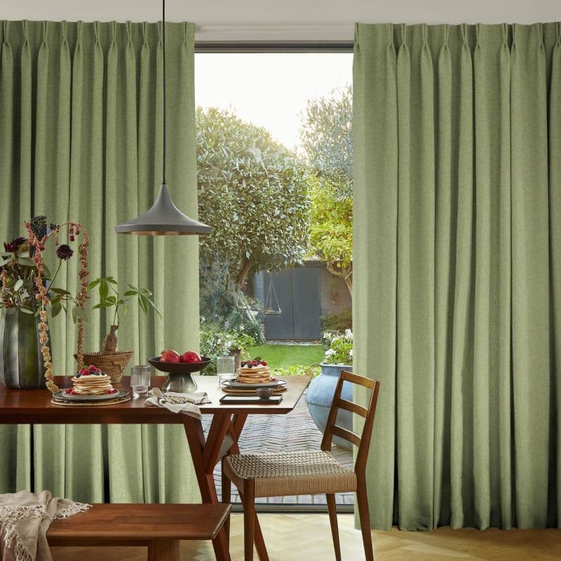 Full-length pleated sage-green curtains, hanging from a ceiling track and partly drawn to frame an opening; filtering daylight into dining area with wooden table, pendant lamp and view of a garden.