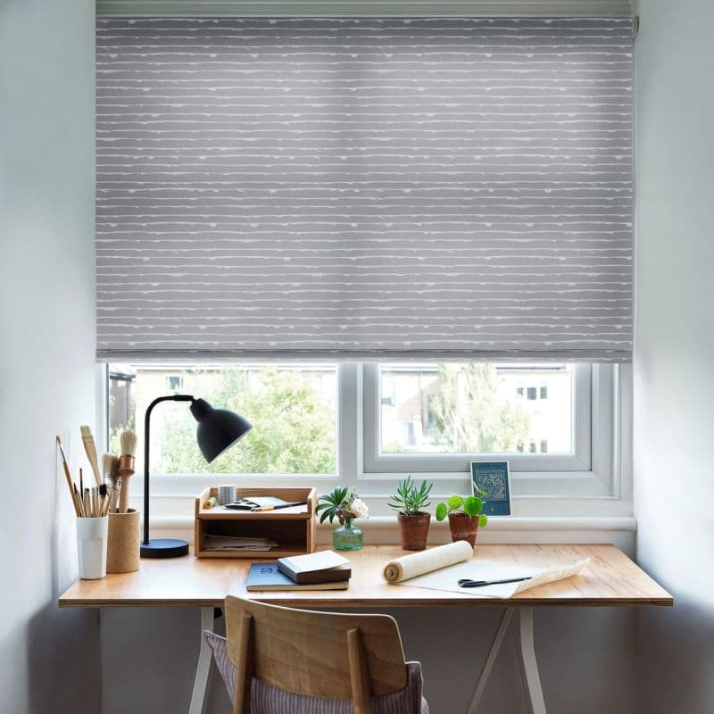 A grey, horizontally striped roller blind is lowered fully, filtering daylight through a small gap at the bottom; beneath it a wooden desk holds a lamp, brushes and potted plants.