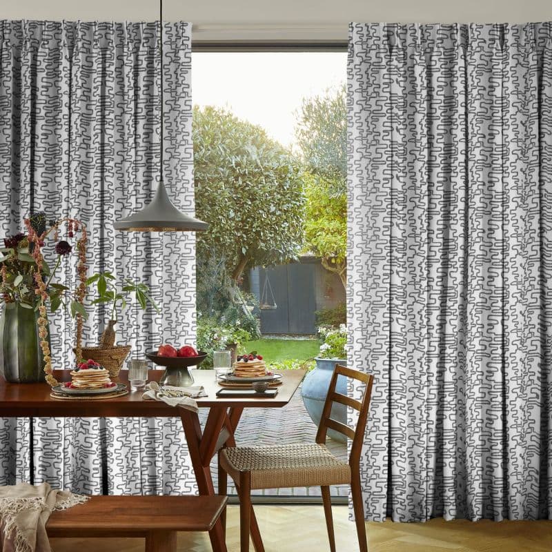 Patterned floor-length pleated curtains (white with gray squiggles) are drawn aside, framing an open patio door; a sunlit dining table set for breakfast overlooks a leafy backyard.