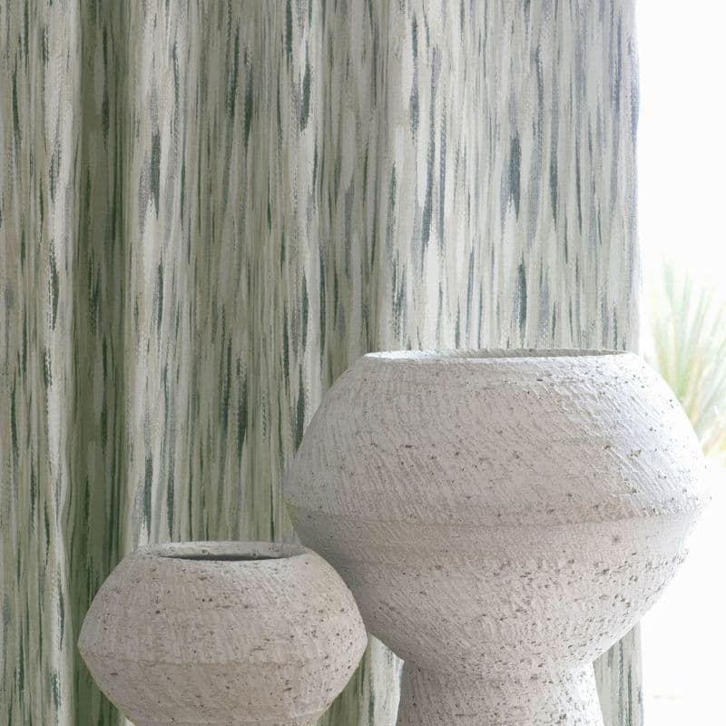 Full-length vertical-striped fabric curtains with green-gray watercolor streaks, drawn closed and gently filtering soft daylight, set behind two textured white stone vases in a softly lit interior near a window.