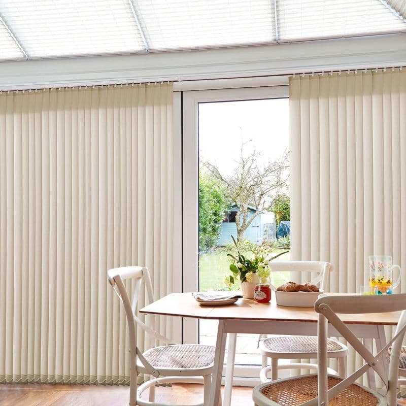 Beige vertical blinds (floor-to-ceiling) across glass patio doors; slats mostly closed, partially drawn revealing central gap, softly filtering daylight. Dining nook with wooden table, chairs, vase and garden view.