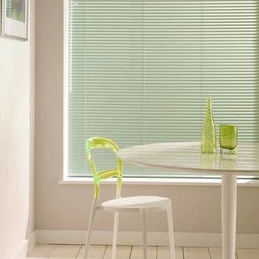 Horizontal pale-green Venetian blinds cover the window, lowered and closed, softly filtering daylight; a dining nook with a white round table, translucent lime-green chair, stool, and light wood floor.