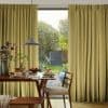 Floor-to-ceiling olive-green pleated curtains, hung and drawn to each side with textured fabric, framing a central opening. They partially reveal daylight and a dining table set for breakfast; garden beyond.