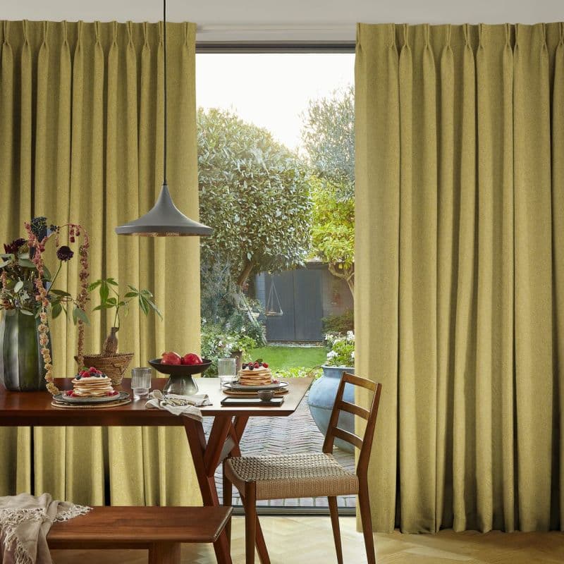 Floor-to-ceiling olive-green pleated curtains, hung and drawn to each side with textured fabric, framing a central opening. They partially reveal daylight and a dining table set for breakfast; garden beyond.