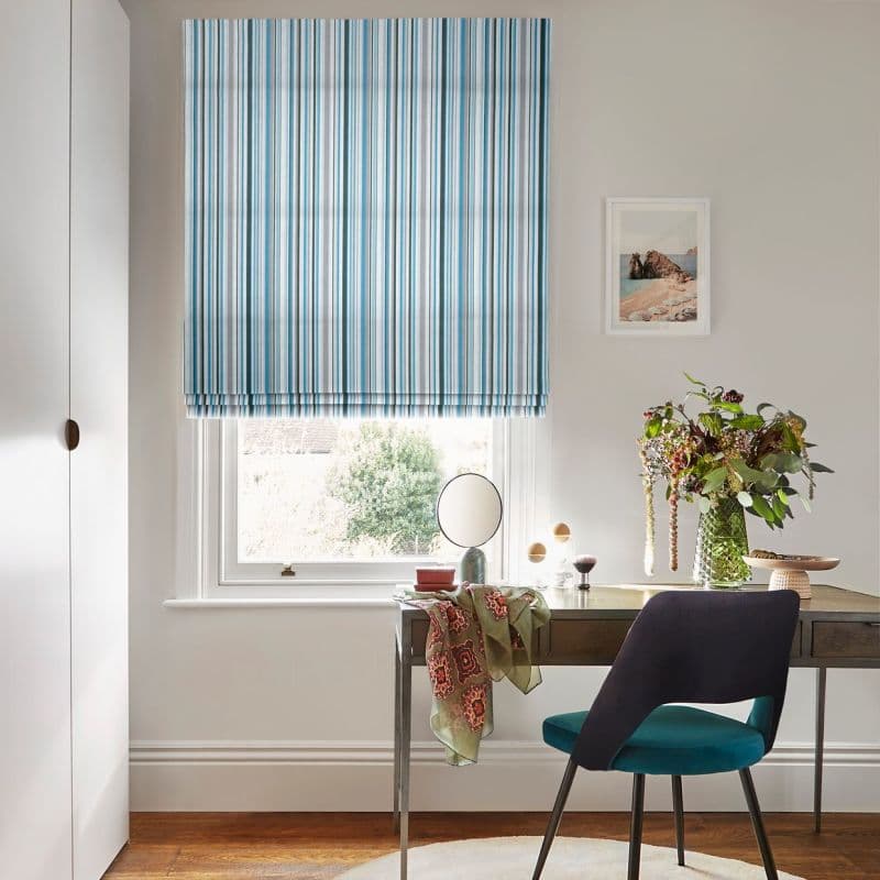 Blue-and-white striped Roman blind, lowered with neat folds, filtering soft daylight into a small home office featuring a wooden desk, teal chair and a vase of flowers.
