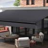 Dark retractable fabric awning with metal roller and valance — fully extended — shading a glass-walled patio furnished with cushioned armchairs, round wooden table and built-in bench beneath brick facade, daylight.