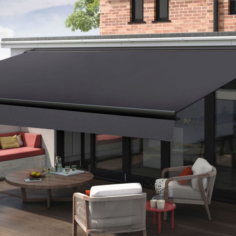Dark retractable fabric awning with metal roller and valance — fully extended — shading a glass-walled patio furnished with cushioned armchairs, round wooden table and built-in bench beneath brick facade, daylight.
