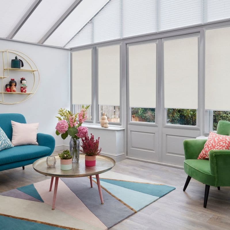 Cream roller blinds on tall French doors and upper windows, partially lowered and softly filtering daylight; bright conservatory with glass roof, teal and green seating and a coffee table.