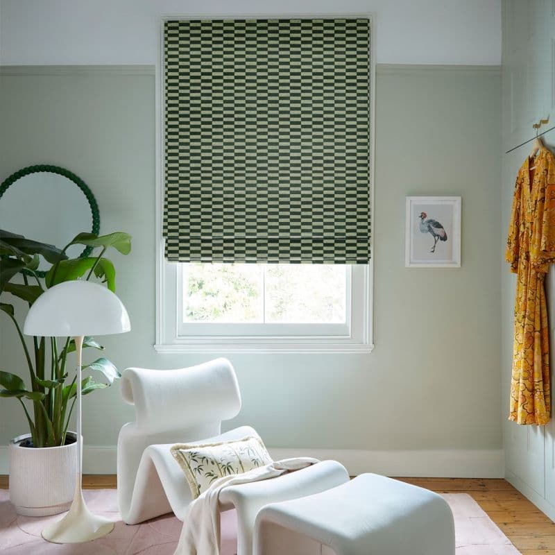 Green-and-beige checkerboard Roman blind lowered over window, fabric showing a slight bottom gap letting light through; in a pale-green sitting nook with white lounge chair, lamp, plant, framed bird print.