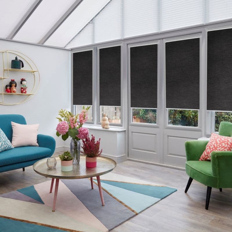 Dark grey roller blinds, lowered across tall conservatory windows and French doors, blocking upper light while leaving lower panes exposed; bright sunroom furnished with teal sofa, green armchair, coffee table.