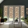 Patterned brown-and-white roller blinds cover French doors, diffusing sunlight and partially blocking the garden view. Modern living room with green armchairs, glass coffee table on a chevron rug.