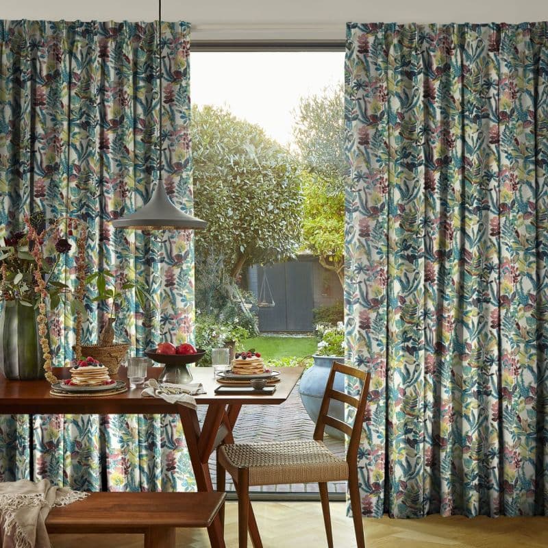 Pleated floor-length floral curtains (teal, pink, yellow) parted centrally, revealing a sliding glass door and admitting soft morning light; wooden breakfast table with pancakes, pendant lamp, woven chair, and garden.