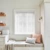 White horizontal slatted blinds, fully lowered and mostly closed, filtering soft daylight; above a built-in cushioned window seat with patterned cushions, a throw, wall sconce and floating shelves in a pale kitchen nook.
BREAD