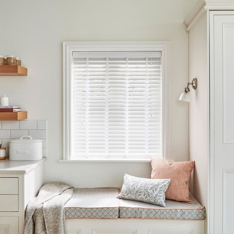 White horizontal slatted blinds, fully lowered and mostly closed, filtering soft daylight; above a built-in cushioned window seat with patterned cushions, a throw, wall sconce and floating shelves in a pale kitchen nook.
BREAD