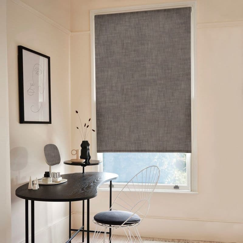 Gray textured roller blind, fully lowered and filtering light; home office with black curved desk, wire chair, minimalist line-art on wall, vase and mirror, soft daylight from lower window.