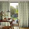 Full-length patterned curtains (green abstract squiggles) on a track, parted centrally; framing a sliding glass door and admitting soft daylight into a dining area set for breakfast with a garden view.