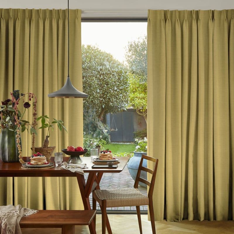 Full-length pleated mustard curtains, drawn mostly closed with a central gap, filtering warm daylight; they frame a dining table and pendant lamp and reveal a sunlit garden beyond.