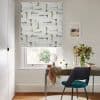 Patterned fabric Roman blind (light gray with gold and charcoal brushstrokes) — partially lowered, filtering daylight — over a desk with teal chair, vase of flowers, pale walls and garden view.