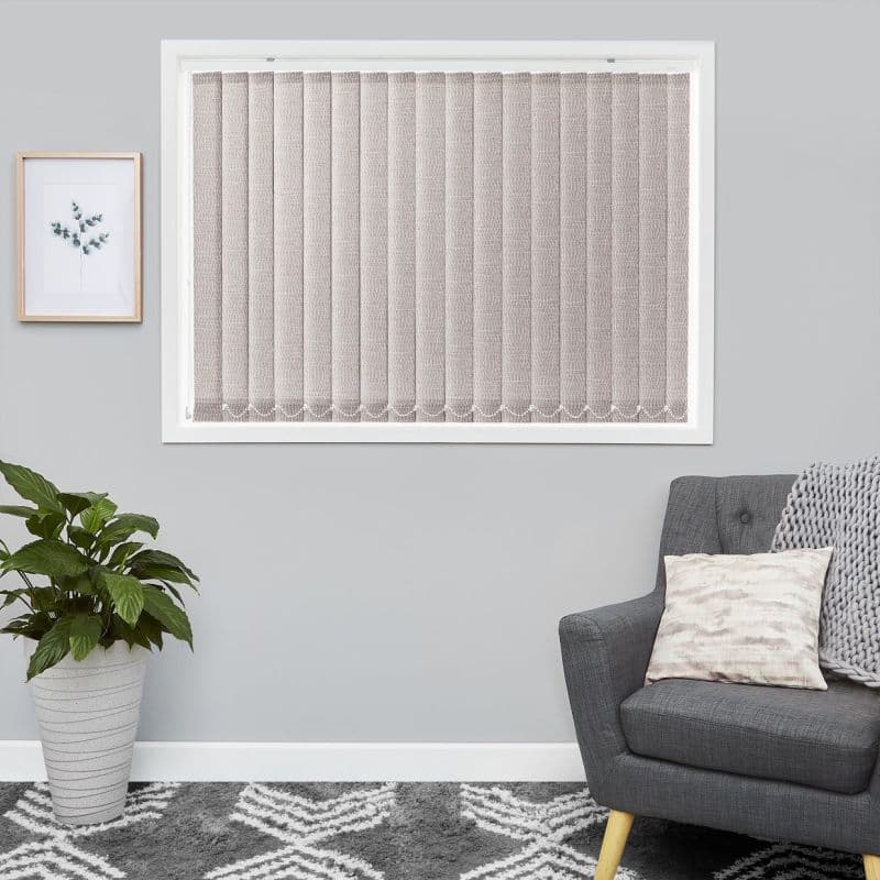 Fabric vertical blinds, beige textured slats fully closed across a white-framed window, gently diffusing light; small living room with grey walls, armchair, knit throw, potted plant and framed art.