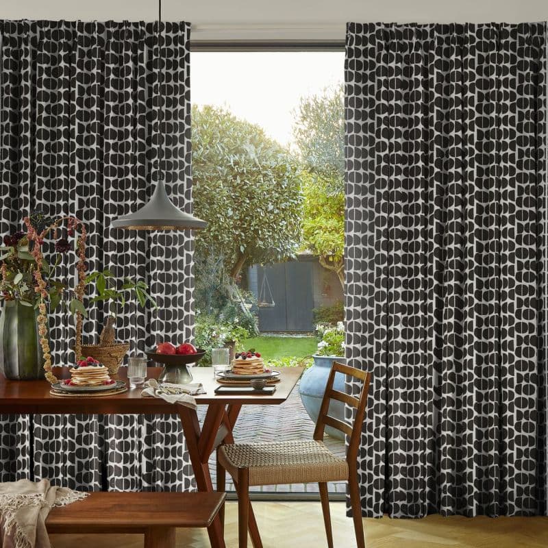 Floor-length black-and-white geometric curtains, drawn partly aside to reveal a glass door; letting morning light into a dining area with wooden table set for breakfast and a leafy garden beyond.