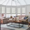 White pleated cellular blinds on conservatory windows and roof, closed and softly diffusing daylight; cozy sunroom living area with beige sofa, chunky pink throw, wooden coffee table on blue rug.