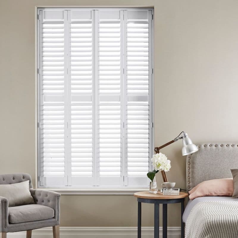 White interior plantation shutters—four hinged panels of horizontal slats—cover the window. They are closed, diffusing bright daylight into a neutral bedroom with an upholstered bed, side-table lamp, and armchair.